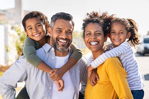 Mixed race parents giving piggyback ride to their children outdoor. Portrait of happy african mother and indian father with daughters looking at camera outside house. Smiling and joyful family standing with their beautiful little girls.